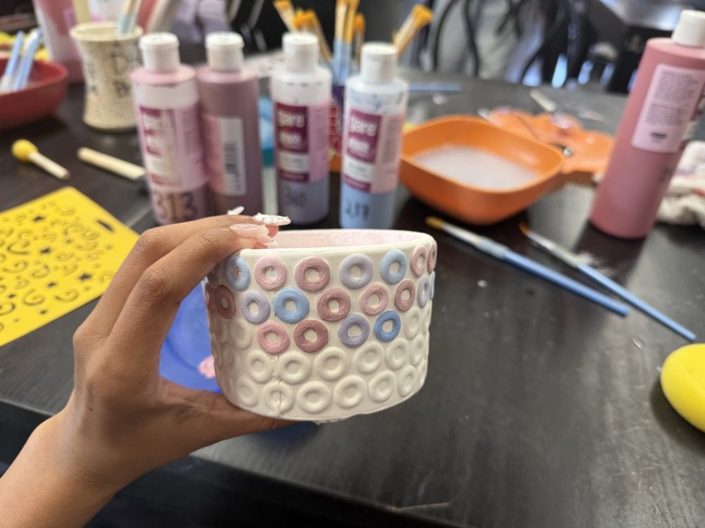 A hand holding a painted ceramic bowl with a ring pattern, paint bottles on the table behind